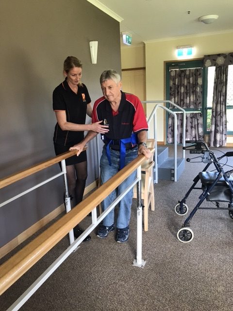 Optimum physiotherapist helping an older client stand tall at parallel bars